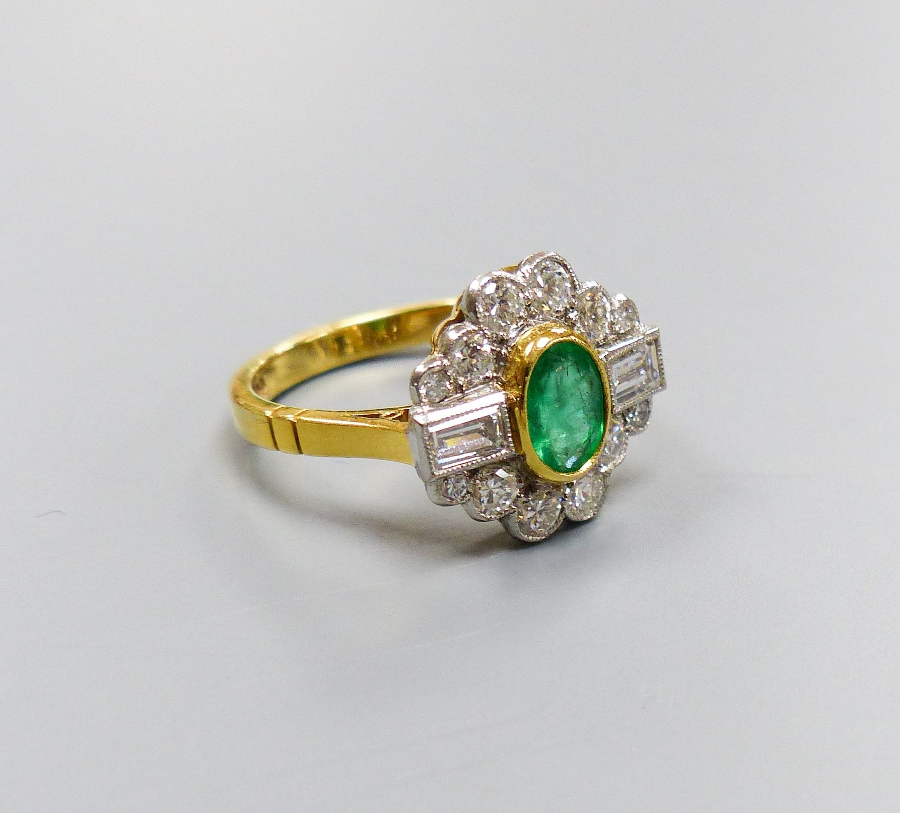 A modern 18ct gold, emerald, baguette and round cut diamond set shaped oval cluster ring, size L/M, gross weight 4.4 grams.